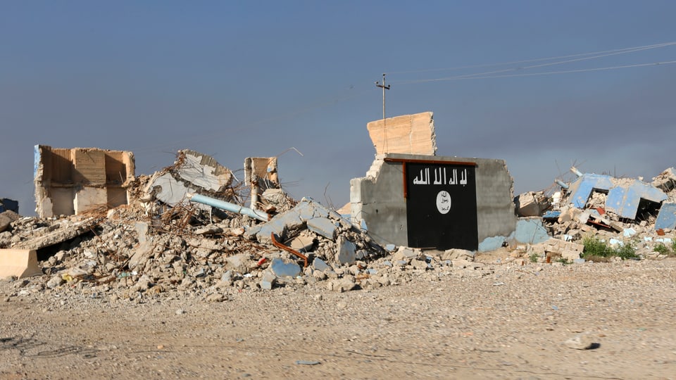 Die Flagge der IS ist auf eine Hausruine gemalt, dahinter sind zerstörte Gebäude sichtbar.