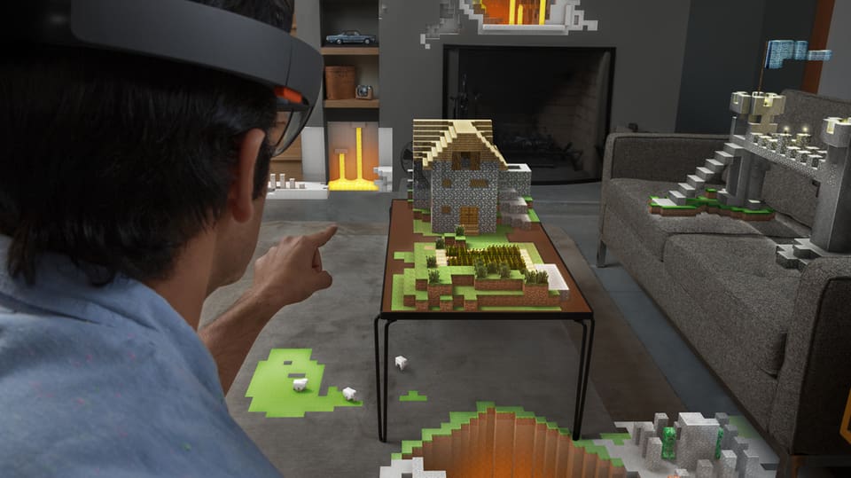 Mann mit HoloLens-Brille zeigt auf einen Tisch. Dort sieht man ein Haus aus Minecraft Bauklötzen.