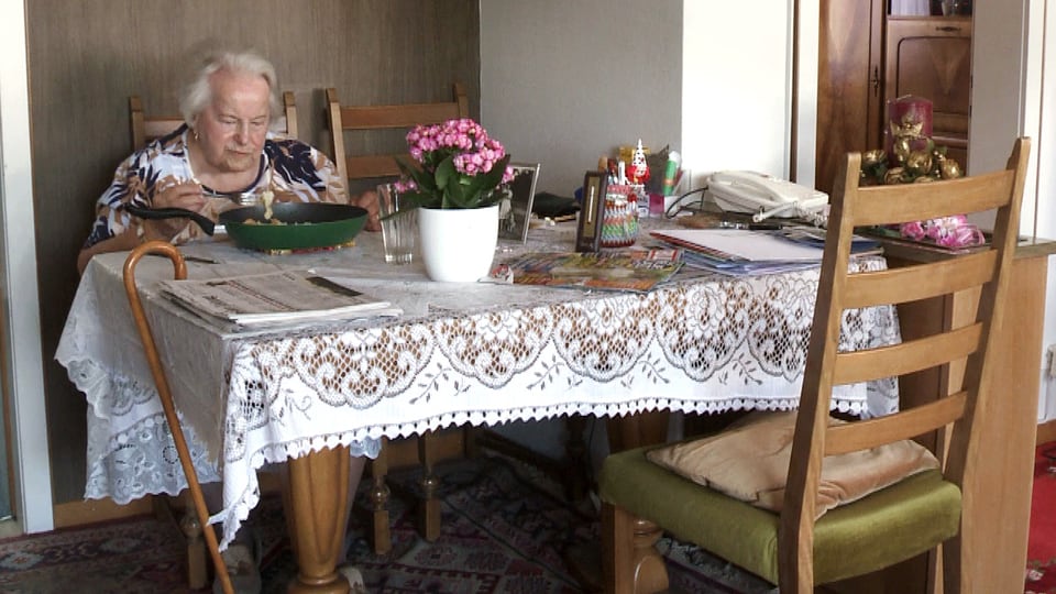 Eine alte Frau sitzt am Tisch mit Häckeldecke darauf, der Stock hängt am Tisch.