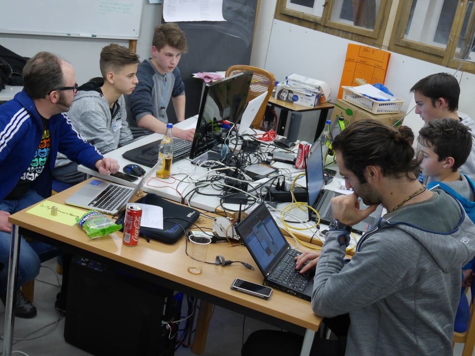 Vier Jugendliche und zwei Erwachsene sitzen an einem Tisch, auf dem Computer und Kabel stehen.