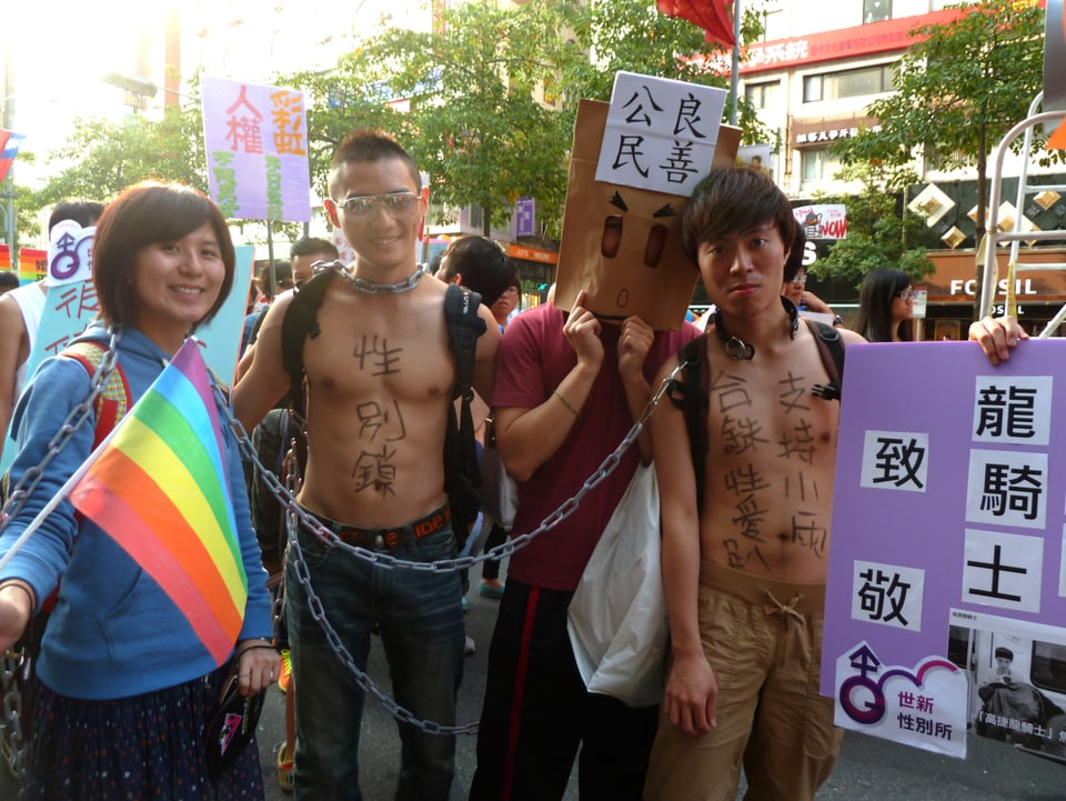 Eine junge Frau und zwei junge Mäner mit nacktem Oberkörper an einer Demonstration.