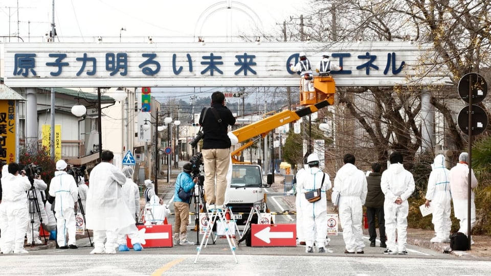 Schild mit japanischem Schriftzug über einer Strasse, darunter zahlreichen Menschen in weissen Anzügen. 