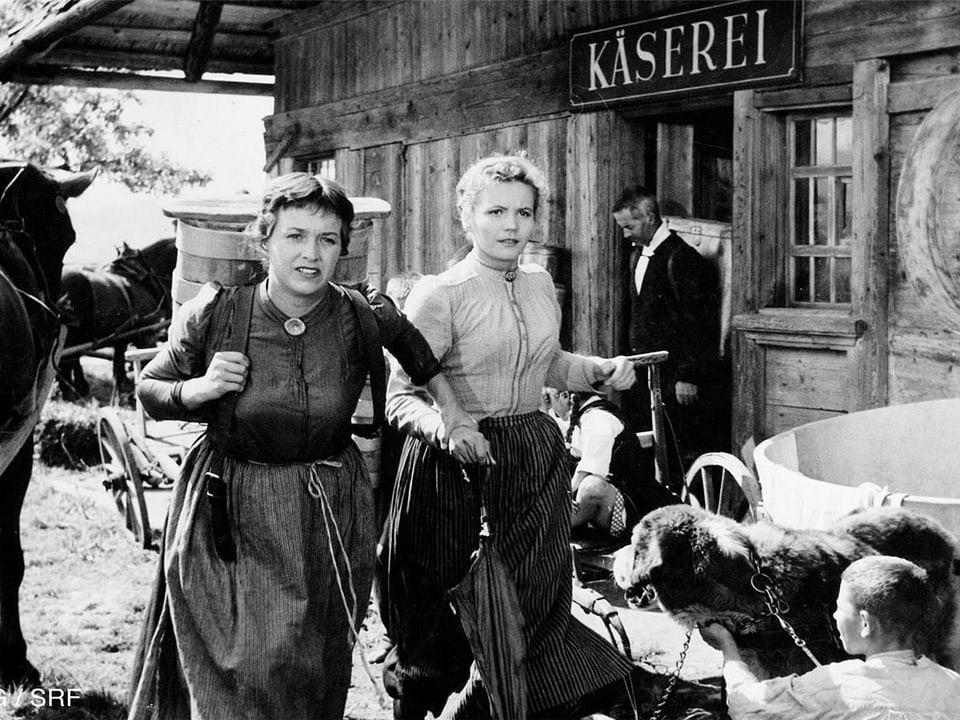 Frauen in Schwarzweiss-Film eilen vor einer Käserei vorbei.