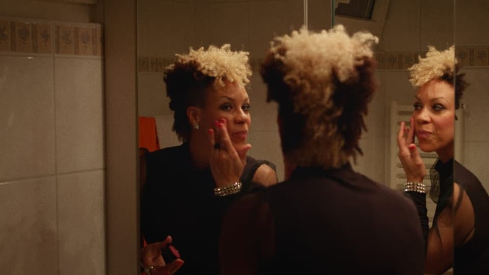 Frau mit kurzen Haaren, unten dunkel oben hell lockig, schminkt sich im Spiegel.