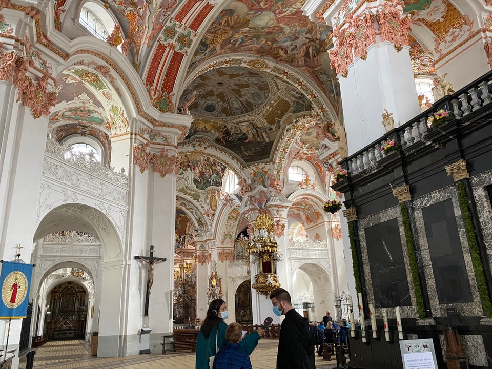 Pater Philipp, Dania und Nick stehen in der Kirche. An der Decke sind üppige und farbenfrohe Verzierungen zu sehen.