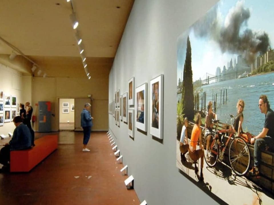 Bild einer Ausstellung, rechts hängt Bild einer Gruppe am Fluss, dahinter Rauch über Gebäude.