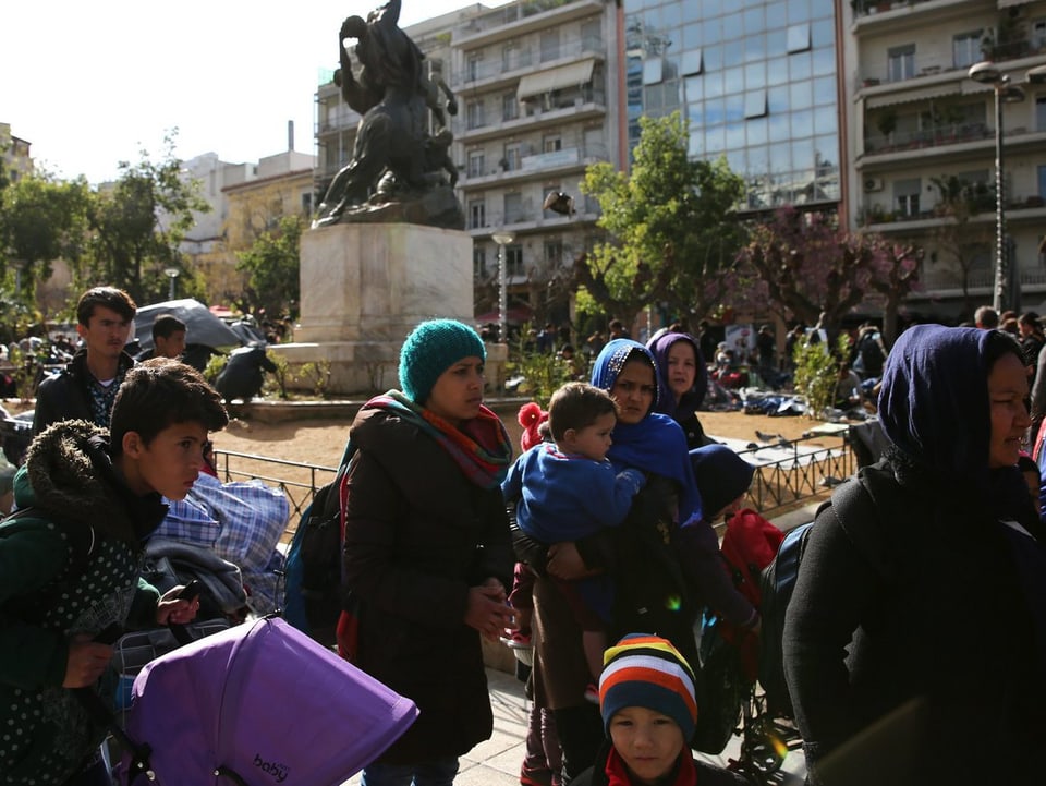 Frauen und Kinder stehen auf einem Platz, im Hintergrund ist eine Statue zu sehen.