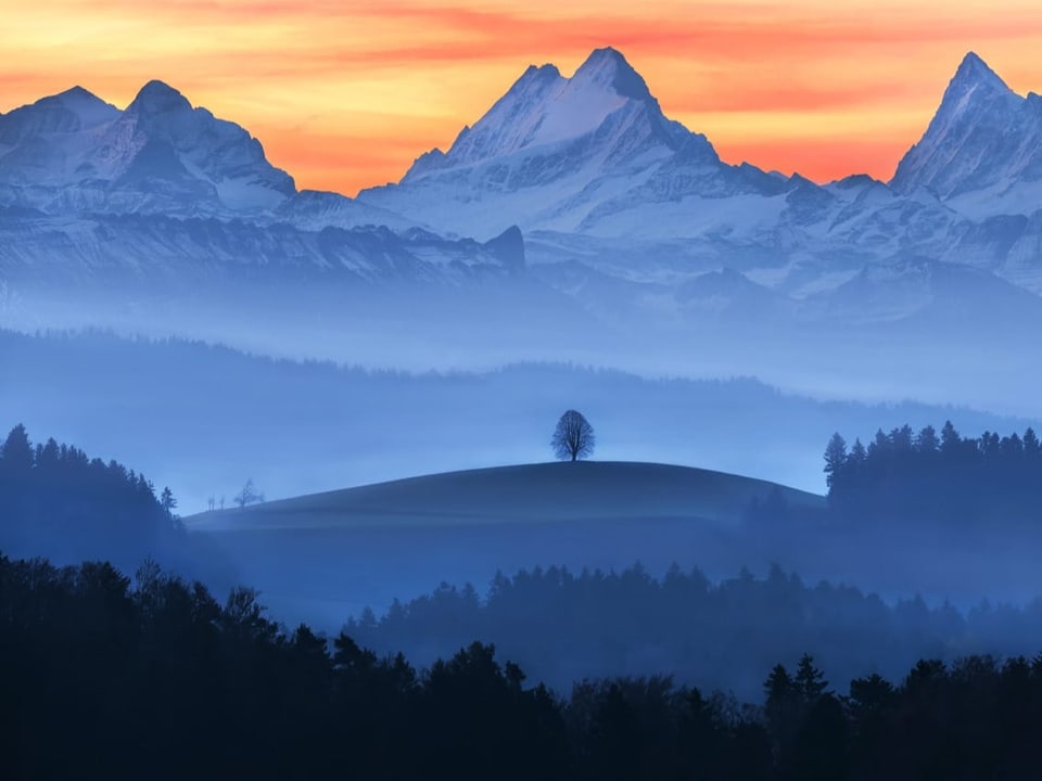 Gelb bis rote Schleierwolken, am Horizont schroffe Schneeberge und im Vordergrund dunstige Täler