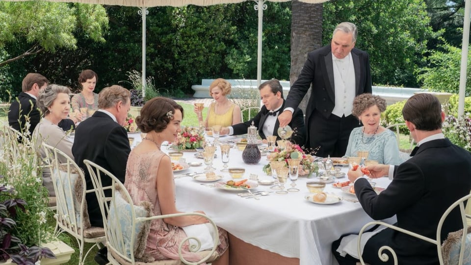 Filmszene: Neun elegant gekleidete Menschen sitzen um einen Gartentisch, ein Mann steht am Tisch und schenkt ein.