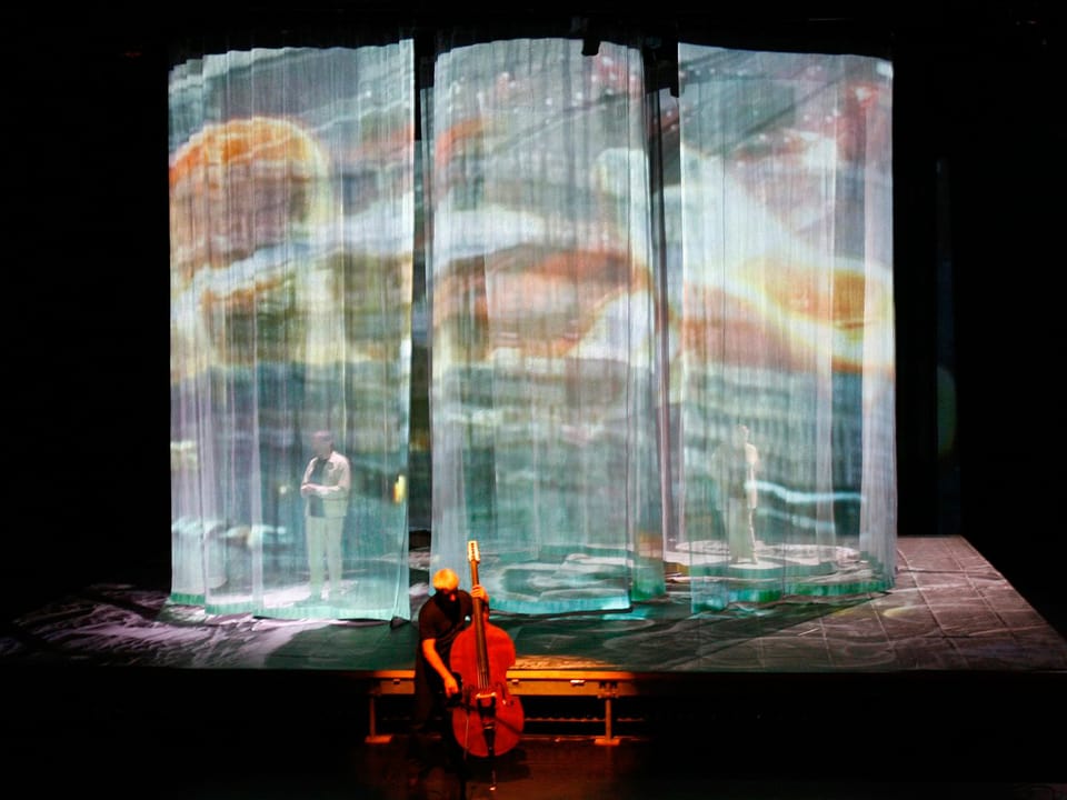 Ein Cello-Spieler steht vor einem Bühnenbild aus halb transparenten Vorhängen.