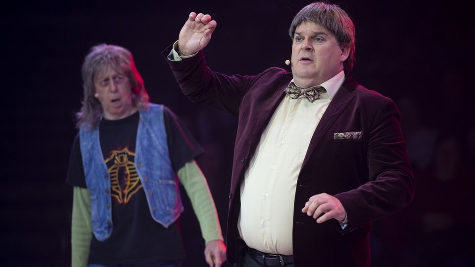 A corpulent man with a bow tie around his neck and a man in a vest stand on a stage and gesture.