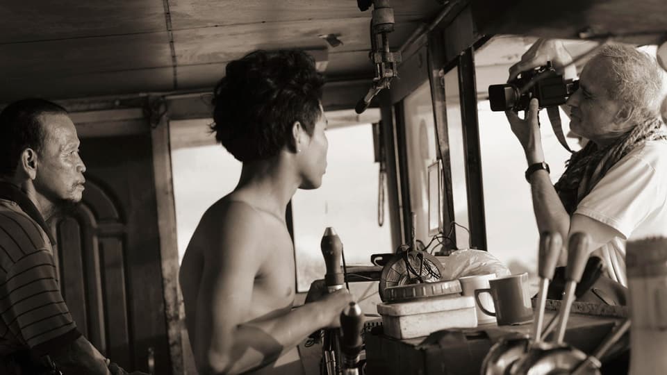 Sepia-Fotografie, ein Mann mit Kamera steht einer Fensterscheibe, hinter der Kamera zwei Männer, einer steuert das Boot