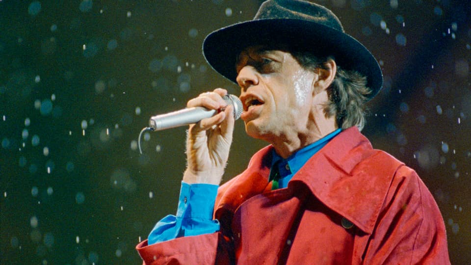 Strömender Regen beim Konzert der Rolling Stones im Basler St. Jakob Stadion. Jagger trägt einen Hut und einen roten Trenchcoat.