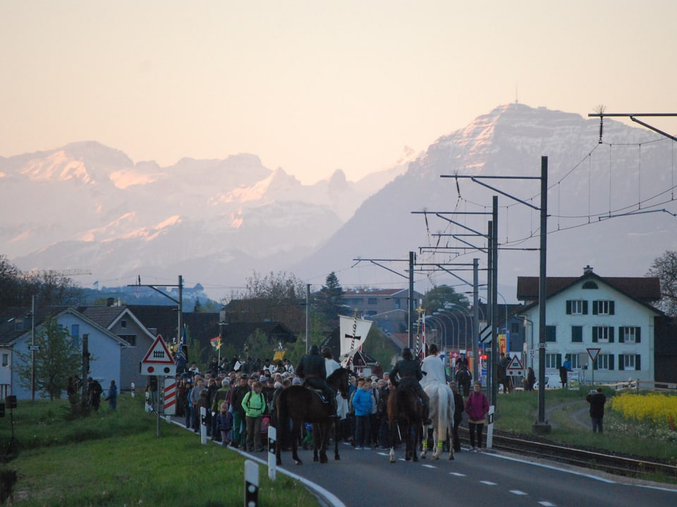 Eine grosse Gruppe von Menschen sammelt sich auf der Strasse, angeführt von Pferden. Im Hintergrund schneebedeckte Berge im Morgenlicht. 