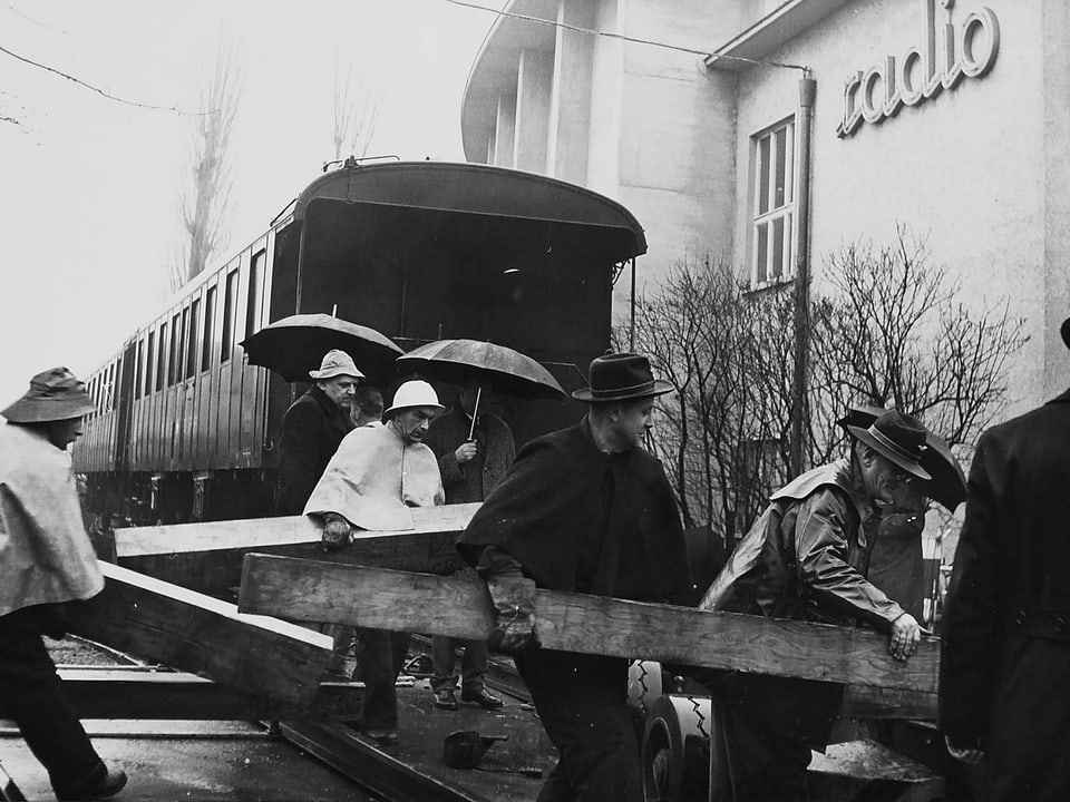 Männer tragen Holzplatten herum. Sie sind vor einem Eisenbahnwaggon auf dem Gelände des Radiostudio Zürich.