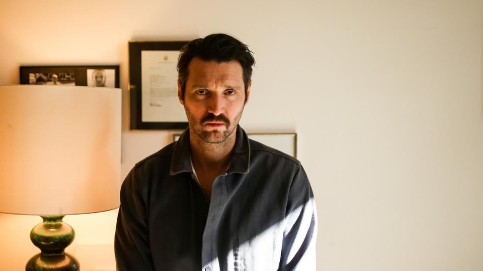 Middle-aged man with black hair and beard, looking blank.