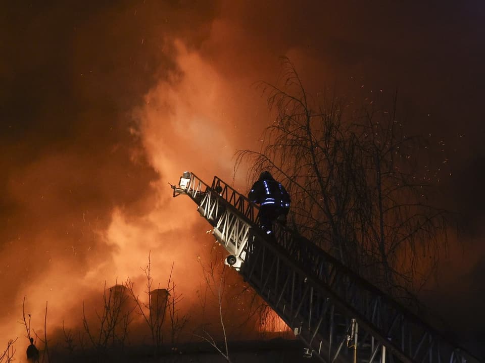 Eine Person in Schutzkleidung steigt auf einer Feuerwehrleiter in das brennende Flammeninferno.