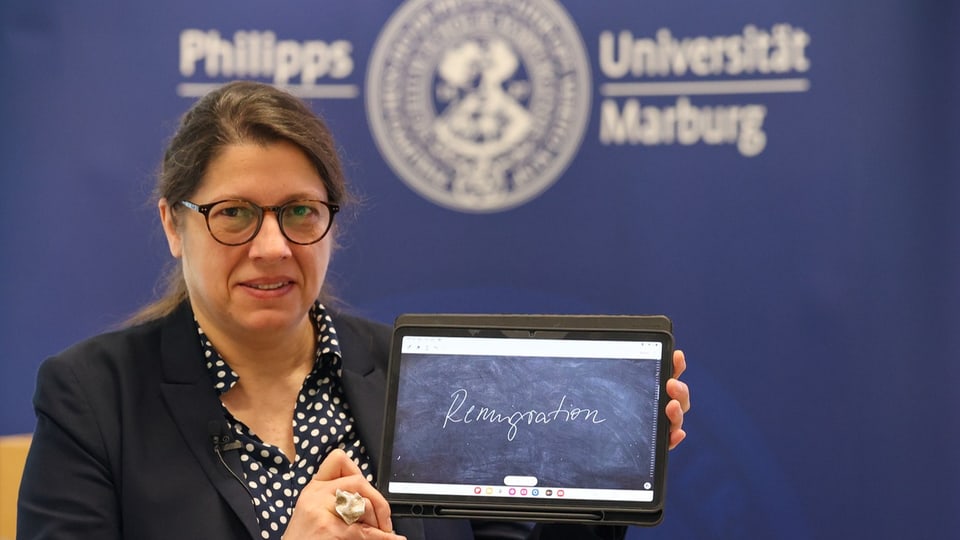Eine Frau Hält ein iPad auf dem das Wort «Remigration» auf einer Tafel steht