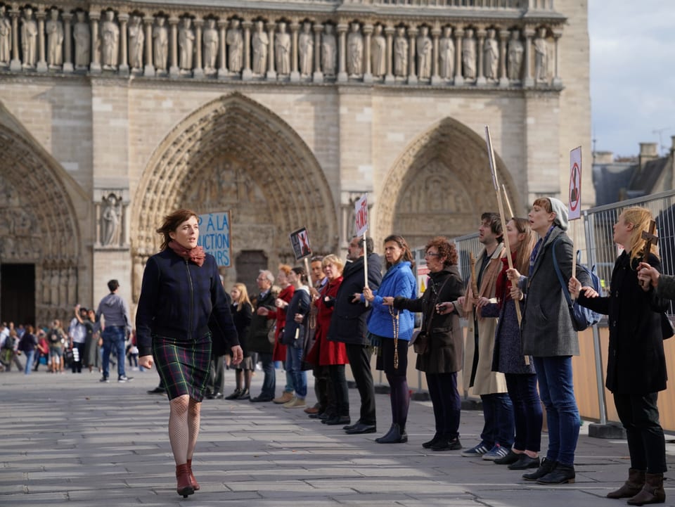Filmszene: Eine frau geht über den Platz vor der Notre Dame. Daneben stehen in einer Reihe Menschen, die Schilder hochhalten. 