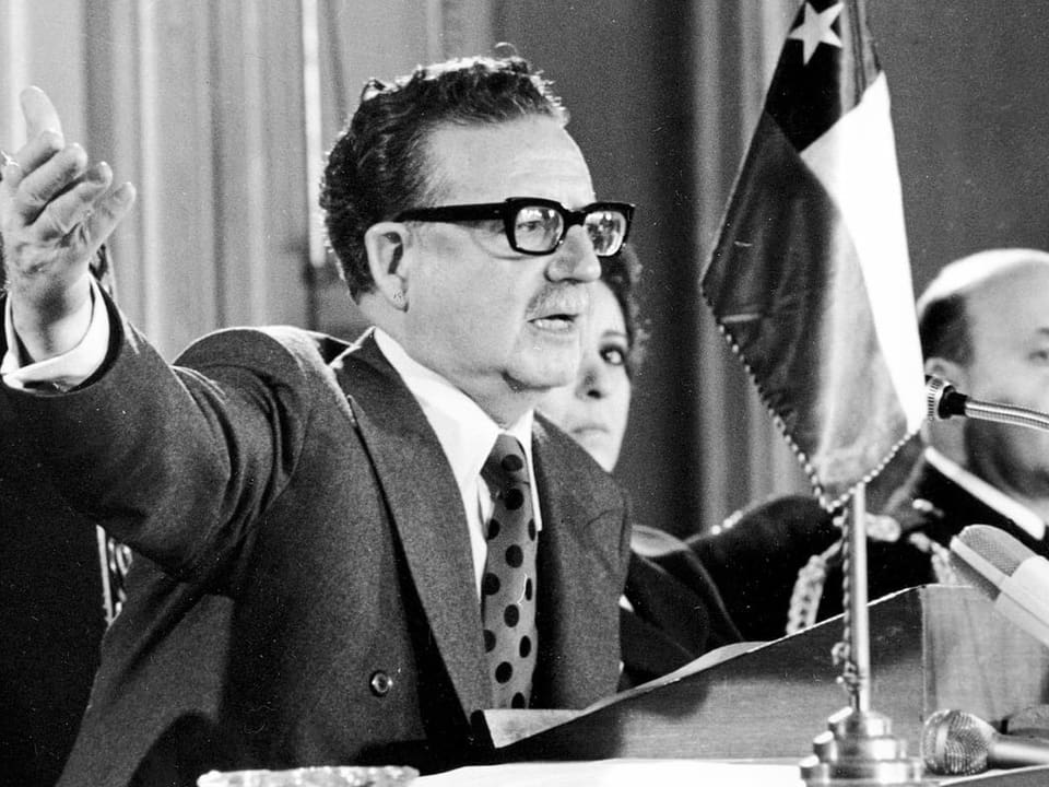 Bild in schwarz/weiss. Salvador Allende spricht an einem Rednerpult und in ein Mikrofon und gestikuliert mit seinem rechten Arm.
