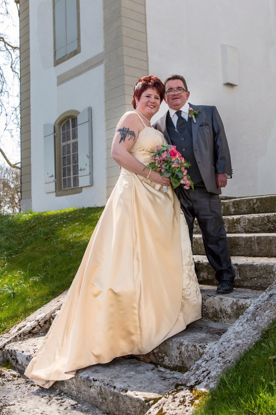 Das Brautpaar, sie im cremefarbenen Kleid, roten Haaren und rosa Blumenstrauss, er im Silvergrauen Anzug, steht nebeneinander auf einer Treppe.
