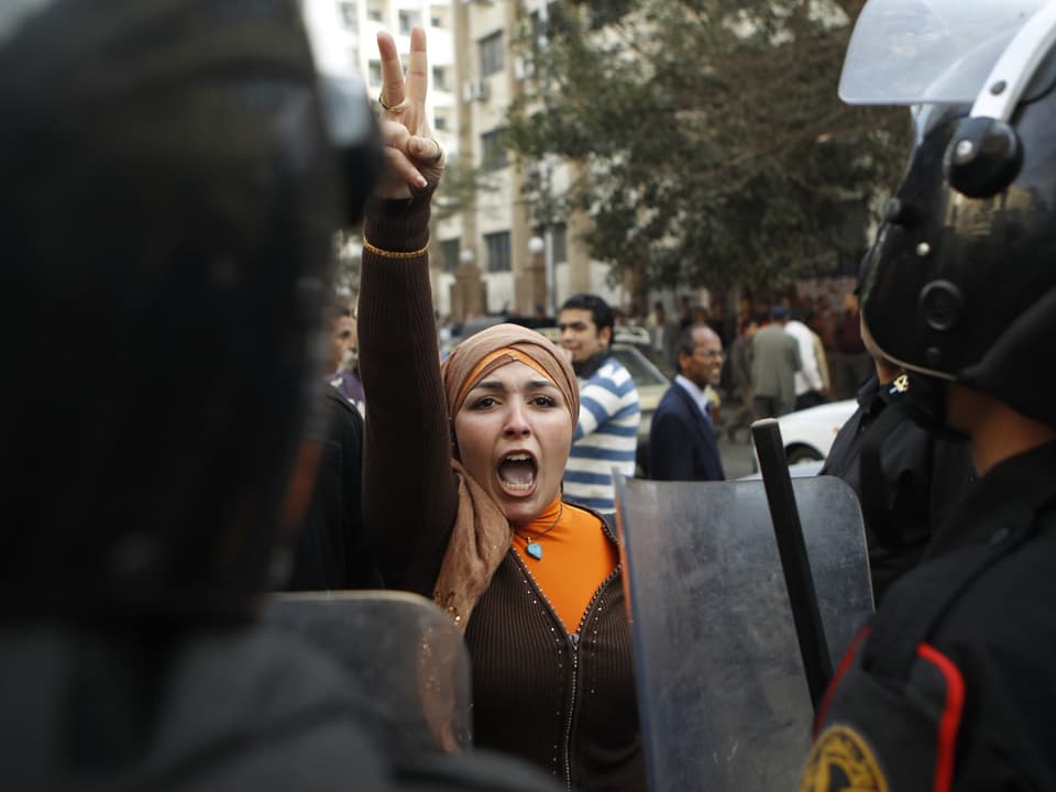 Eine Demonstrantin mit Kopftuch steht vor der Reihe aus Polizisten und zeigt das Victory-Zeichen.