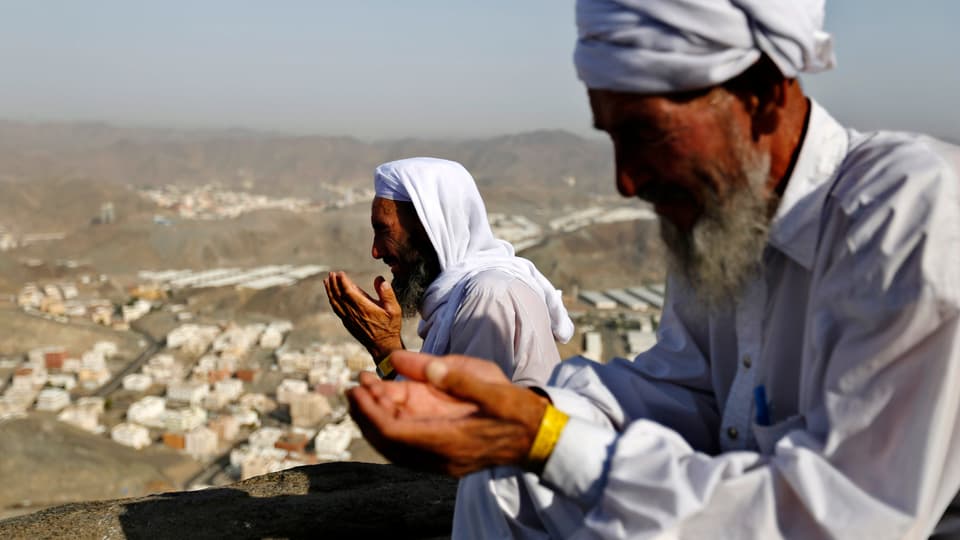 Zwei weiss gekleidete Männer beten auf einem Berg. Im Hintergrund ist die Stadt Mekka zu sehen.