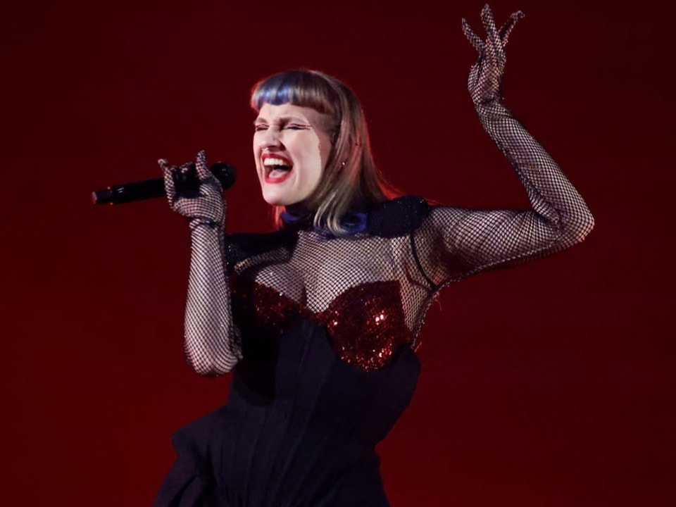 Sängerin in schwarzem Outfit mit Mikrofon auf roter Bühne.