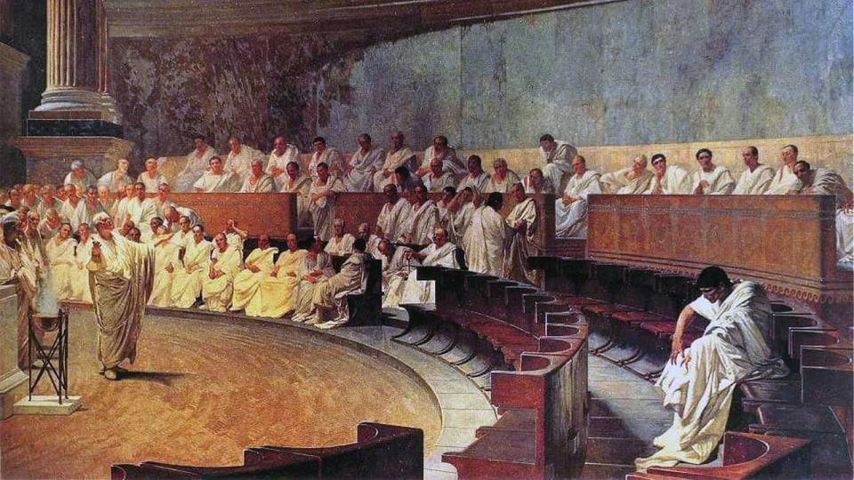 Weiss gekleidete Römer sitzen in einem Saal. In der Mitte spricht eine stehende Person mit ausgebreiteten Armen.