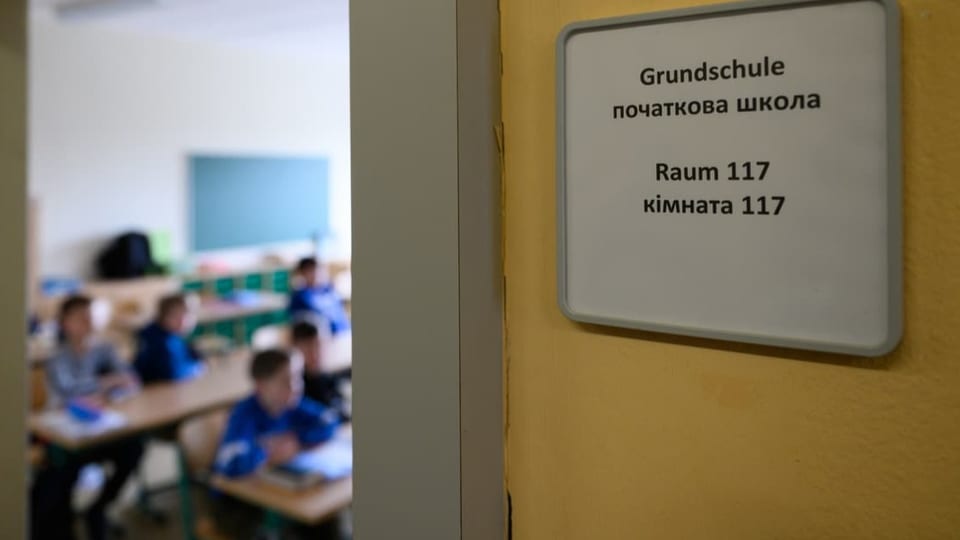 Raum ist angeschrieben mit «Grundschule» und daneben in kyrillischer Schrift. Im Hintergrund sind Kinder an Tischen.