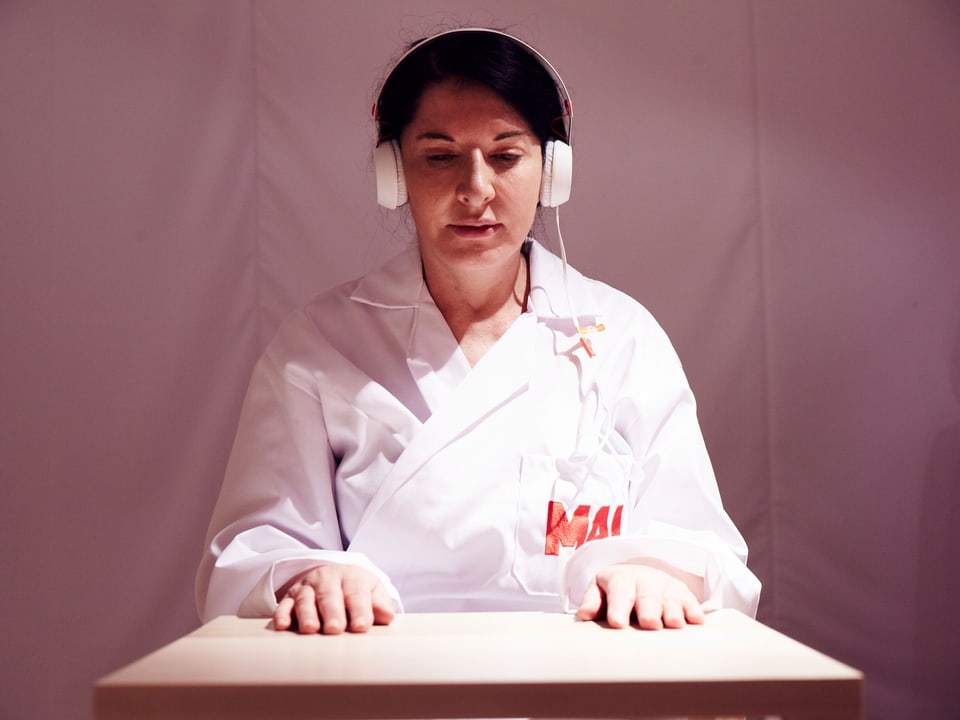 Marina Abramović sitzt in einem weissen Overall mit der Aufschrift "MAI" an einem Tisch, mit weissem Kopfhörer, die Arme auf den Tisch gelegt, 