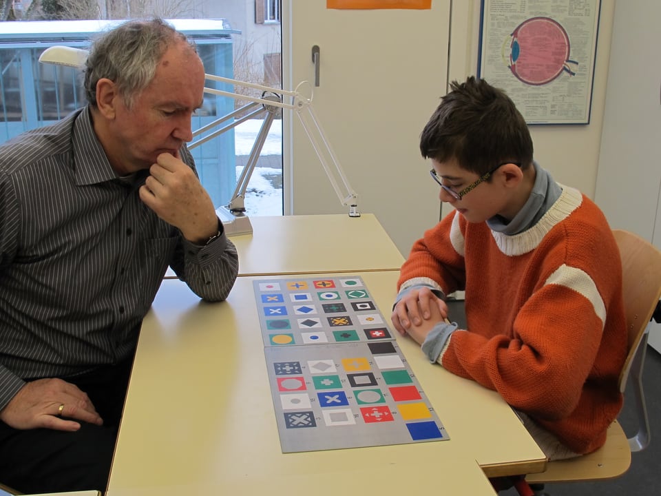 Lehrer Ueli Rüegg und Schüler Vinzenz sitzen sich an einem Tisch gegenüber und betrachten Karten mit unterschiedlichen Formen und Farben darauf.