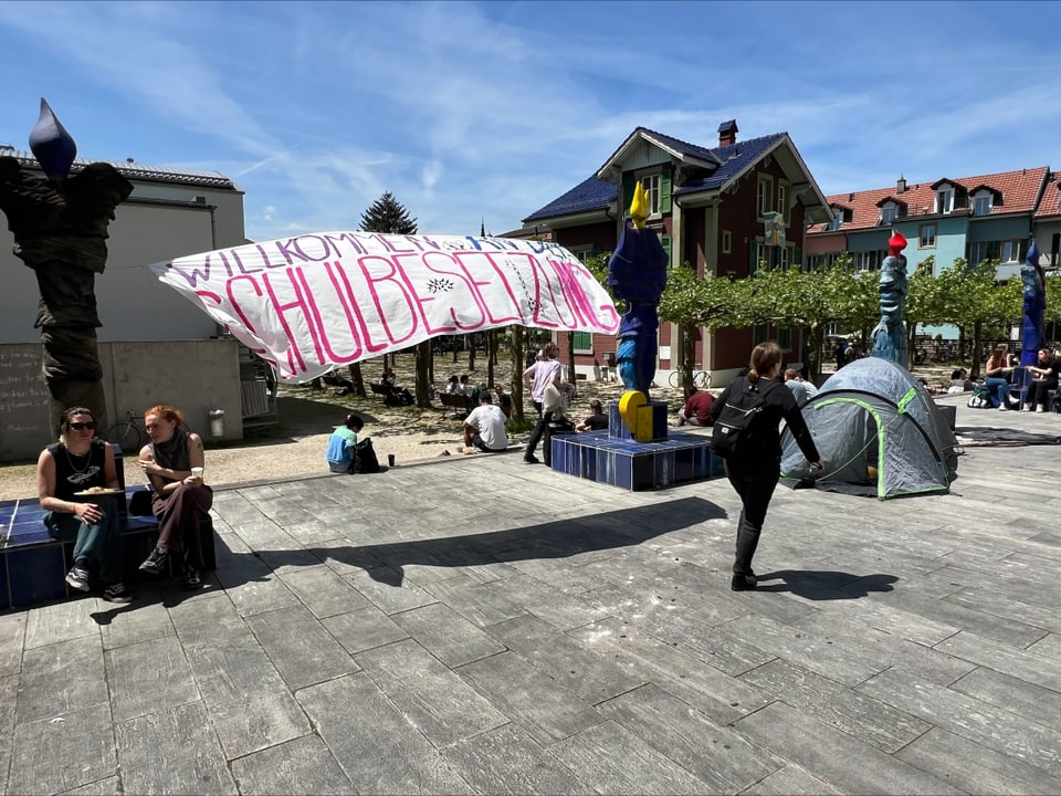 Menschen auf einem Platz mit einem beschrifteten Banner und Zelten.