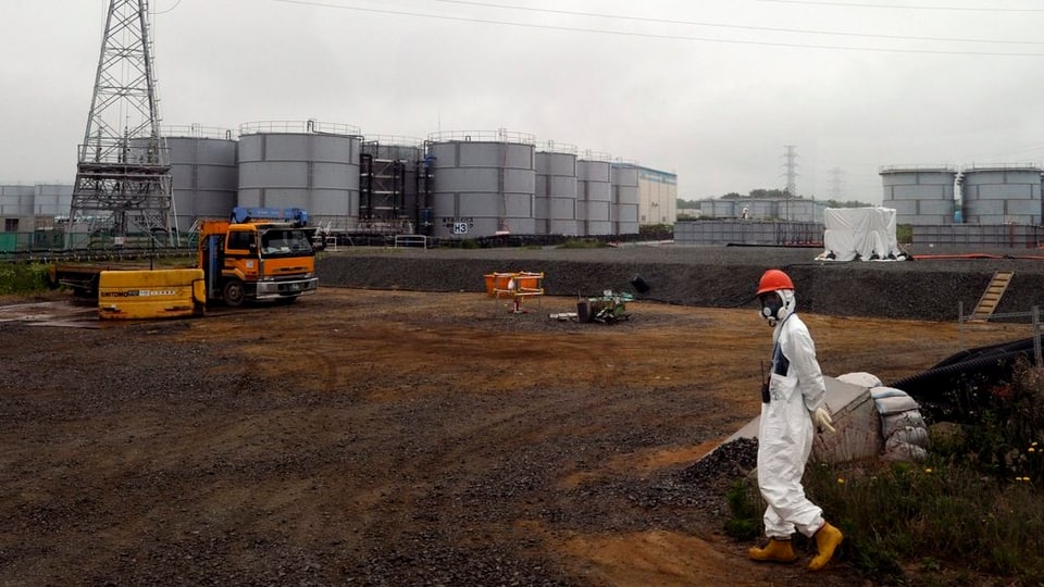 Tanks bei der Atomruine von Fukushima, davor ein Mann in weisser Schutzkleidung.