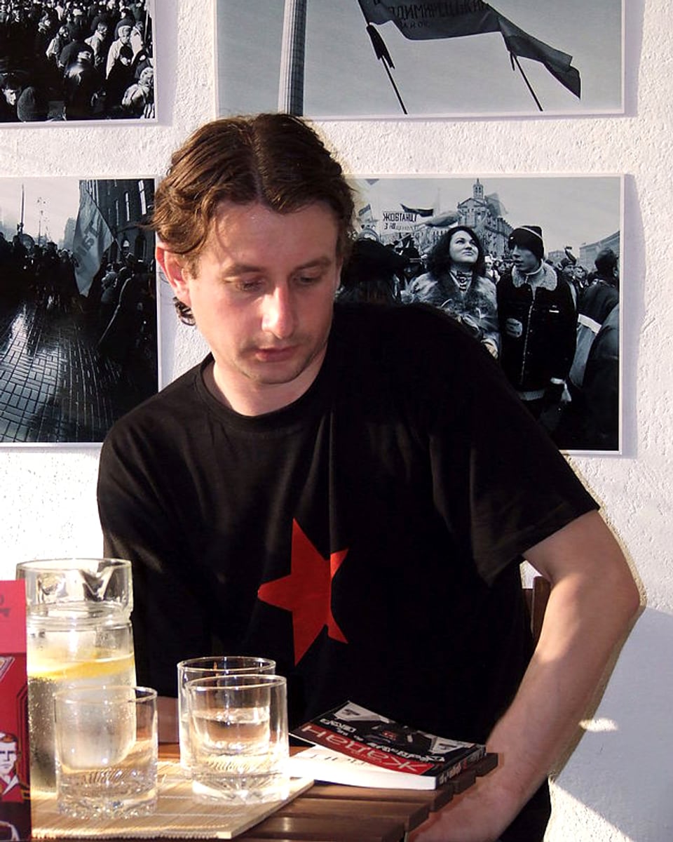 Ein Mann sitzt an einem Tisch, vor sich ein Buch, auf das er blickt. Er trägt ein schwarzes T-Shirt mit einem grossen roten Stern auf der Brust.