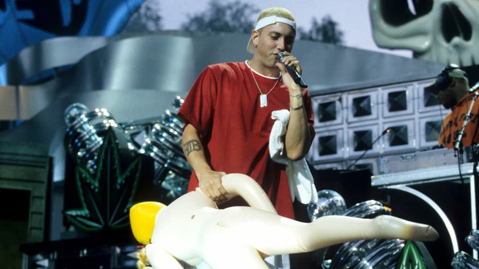 Ein Sänger hält auf einer Bühne eine Gummipuppe in der Hand.
