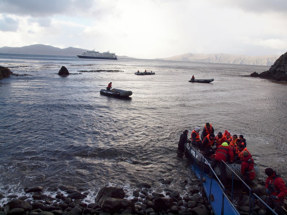 Boote auf dem Meer, im Hintergrund ein Kruezfahrtschiff, im Vordergrund steigen Menschen mit Schwimmwesten an Land. 