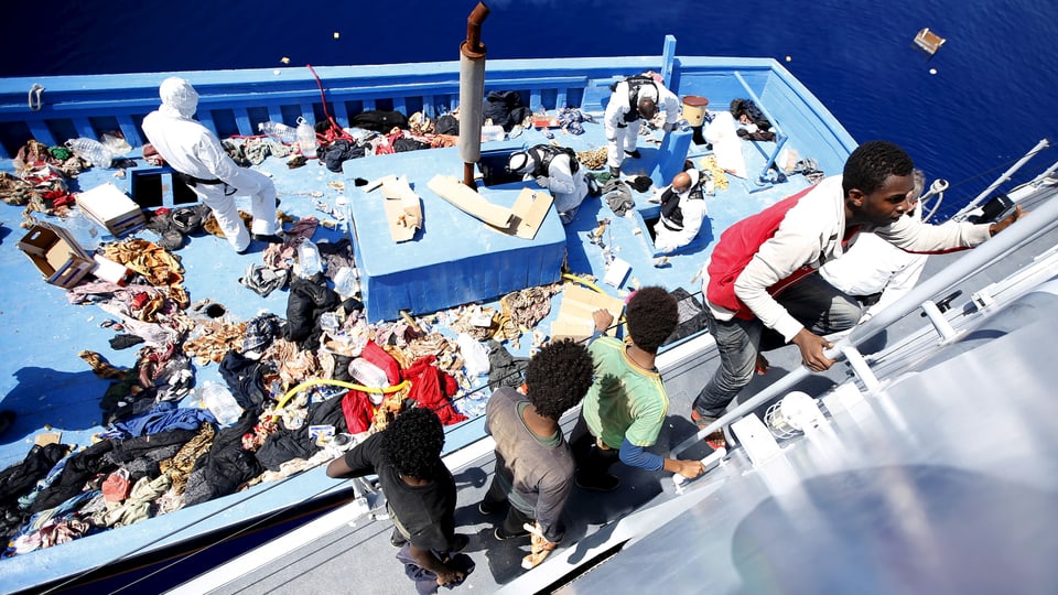 Flüchtlinge steigen die Leiter hoch. Das Deck des Flüchtlingsschiffs ist übersät mit Kleider und Abfällen.