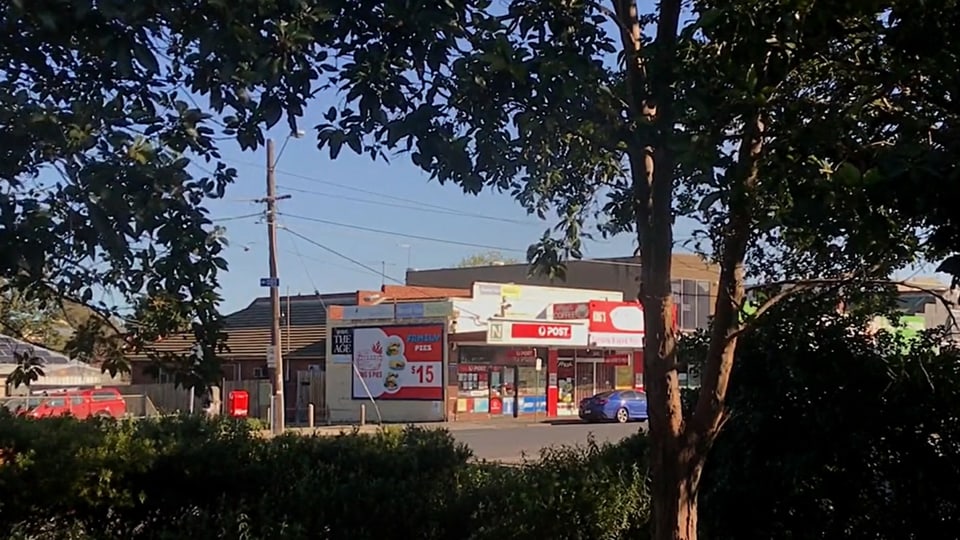 Blick aus einem Fenster. Man sieht eine Strasse und ein kleines Postbüro zwischen dunklen Bäumen hindurch.