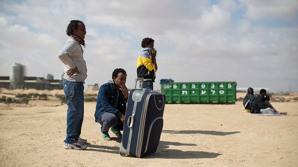 Männer stehen und sitzen in der Wüste, neben ihnen ein grosser Reisekoffer.