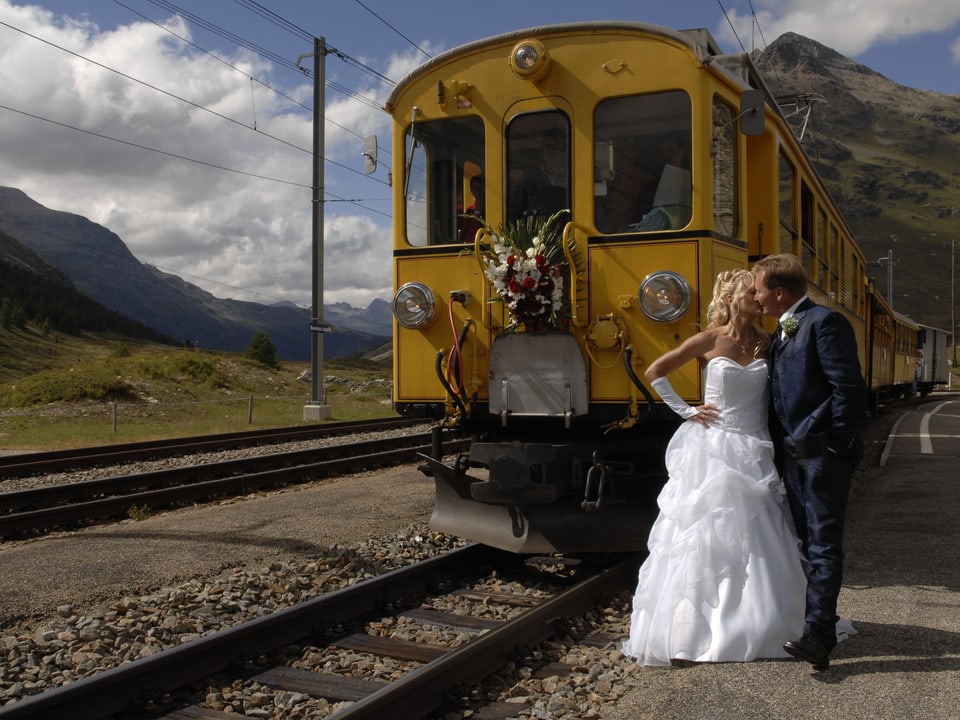 Ein Brautpaar steht küssend vor einem mit Blumen geschmückten, gelben Zugwaggon. Im Hintergrund sieht man Bergpanorama und Wiesen.