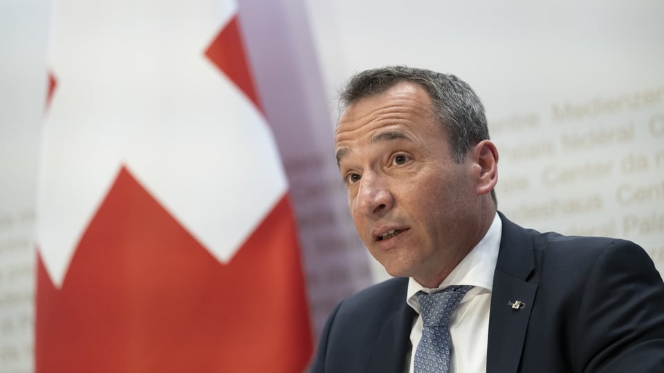 Mann im Anzug spricht bei einer Pressekonferenz vor einer Schweizer Flagge.