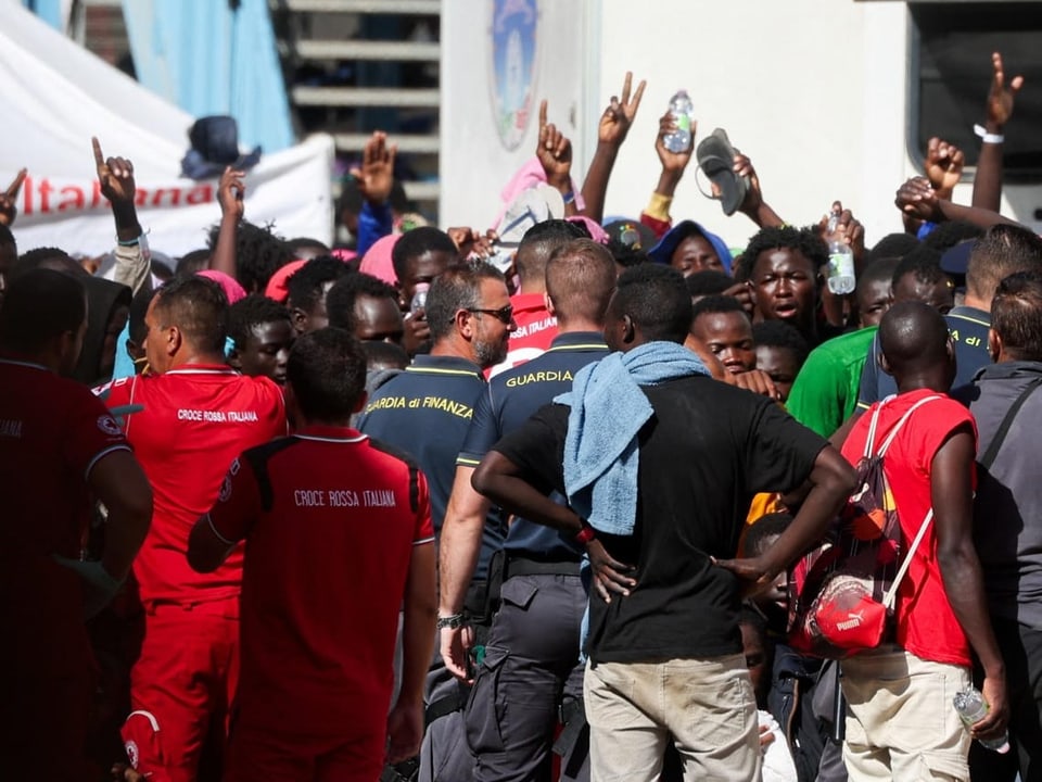 Migranten winken und heben die Hände, Sicherheitspersonal und Rotes-Kreuz-Mitarbeiter