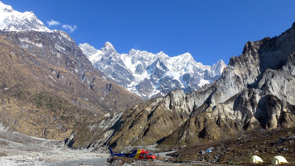 Ein Hubschrauber und zwei Zelte vor einer mächtigen, aber gletscherlosen Bergkulisse im Himalaja.
