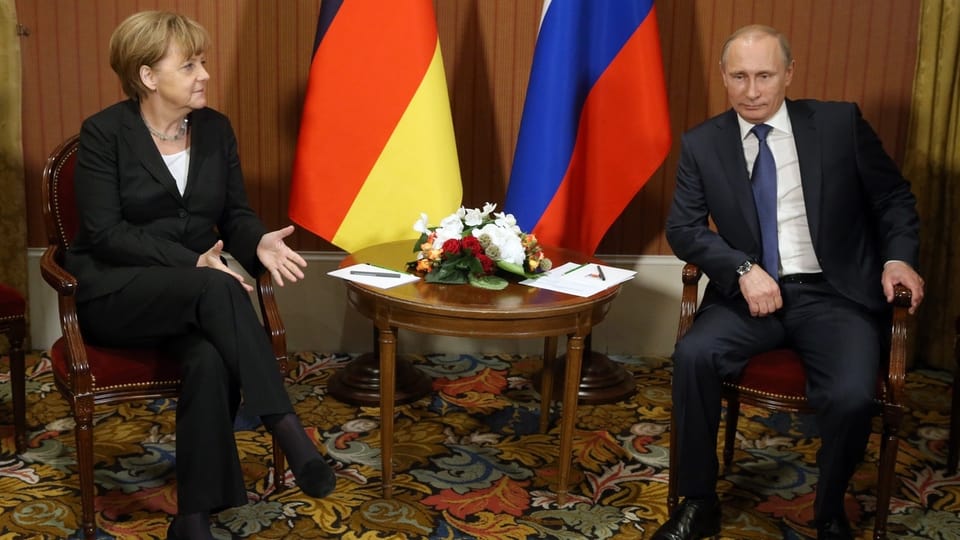Angela Merkel (l.) sitzt neben Wladimir Putin vor Fahnen von Deutschland und Russland