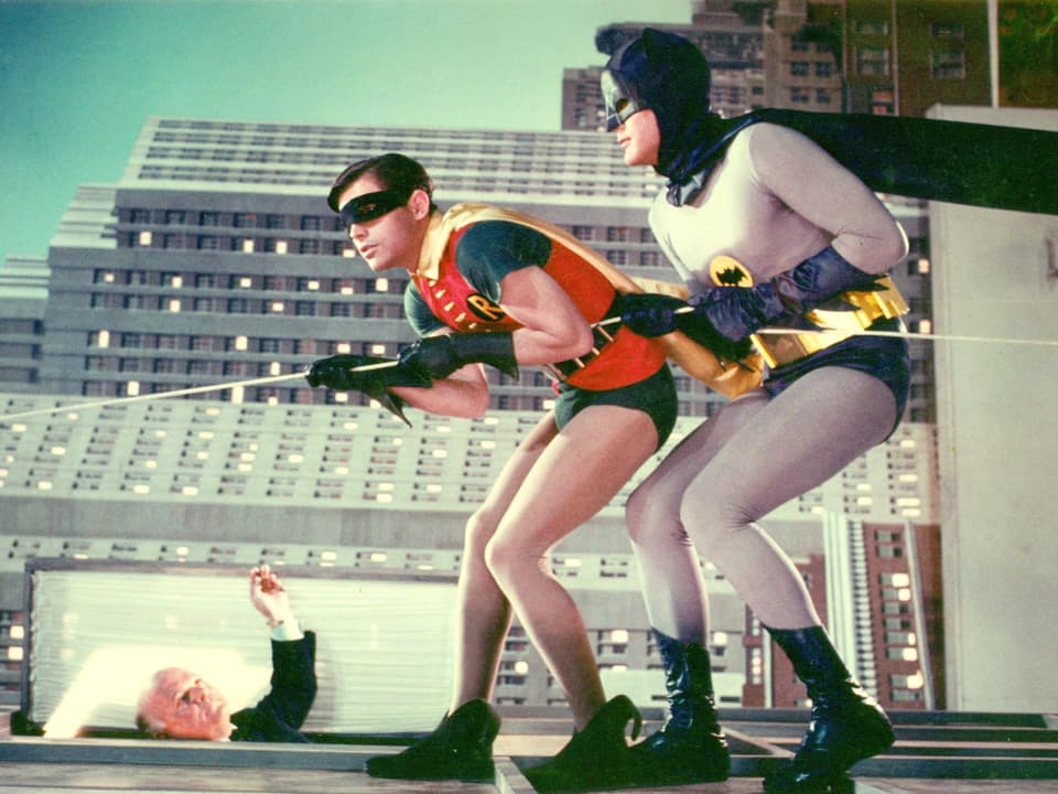 Szene aus der TV-Serie: Batman und Robin, zwei Männer in Superheldenkostümen, klettern an einem Seil eine Gebäudefassade hoch.