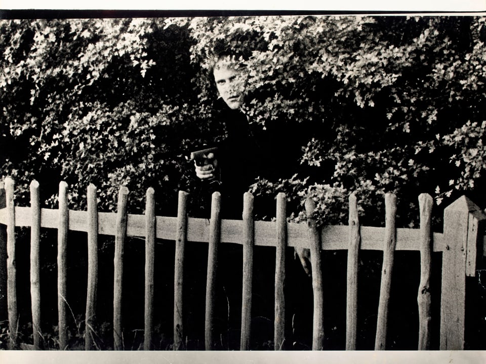 Ein Mann hinter einem Zaun mit einem Gewehrt. Er ist von Gebüsch umgeben.