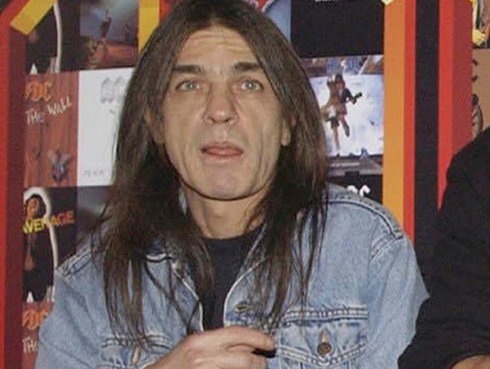 Malcolm Young in Jeansjacke vor einer Bilderwand mit AC/DC-Covern