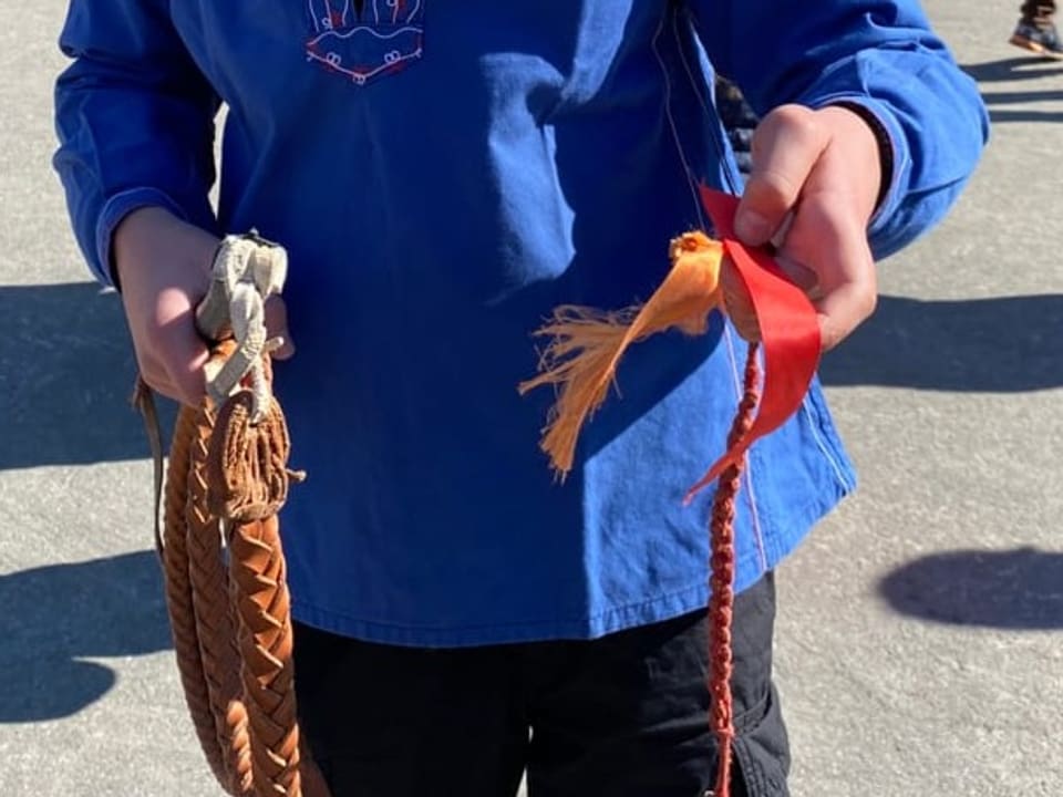 Der «Schnipp» 2 Hände halten eine Peitsche in der Hand. Die Peitsche ist braun, der Zipfel ist orange.