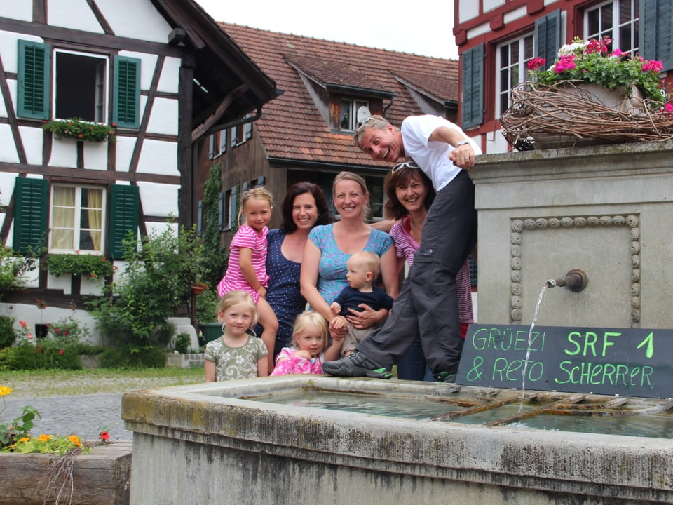 Gruppenbild vor dem Dorfbrunnen in Guntalingen. Reto Scherrer steht auf dem Brunnen, die Frauen mit ihren Kindern davor. Auf dem Brunnen steht ein Willkommensschild.